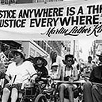 Black and white photo of a 1993 disability rights march in New York City. At front are Paul Miller, Judy Heumann, Justin Dart, and others, with large crowd behind. Marchers holding a sign that says: "Injustice anywhere is a threat to justice everywhere.” Photo credit: Tom Olin.
