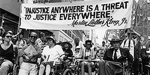 Black and white photo of a 1993 disability rights march in New York City. At front are Paul Miller, Judy Heumann, Justin Dart, and others, with large crowd behind. Marchers holding a sign that says: "Injustice anywhere is a threat to justice everywhere.” Photo credit: Tom Olin.
