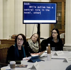 Dr. Brenda Bruggerman is shown seated at a conference, along with two other attendees. All seem to be listening to someone out of the frame and appear female-presenting with light skin. Behind the people is a large TV screen with CART captions reading "... book. While autism has a central role in the novel, other..."