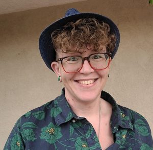 Griffin is seen from the shoulders up in masculine presentation, posed smiling with a blue hat, curly short hair, glasses, and a casual blue and green floral print button down shirt.