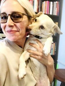 Image Description: A light-skinned woman with white hair and glasses, smiles warmly as she stands before a bookshelf. In one hand, she holds a small white dog, who licks her face.