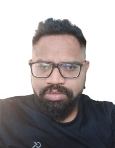 Image Description: A brown-skinned man with short hair and a beard is shown against a white background. His face is neutral, thoughtful. He wears glasses and a black shirt.