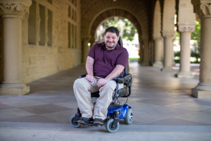 Image Description: A light-skinned man smiles, seated in a wheelchair before a corridor of pillars. He wears khakis and a collared, short-sleeve purple shirt.