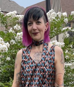 Image Description: A white woman with black bangs and dark pink shoulder length hair. She is wearing a pink, black, and blue geometric satin tank with a thin scarf in the same pattern around her neck. She has various tattoos on her arms and chest. A crepe myrtle tree with white blossoms is behind her. She is smiling with her mouth closed.