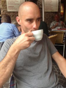 Image Description: A bald white male in a grey t-shirt drinking an espresso, seated in an outdoor cafe.