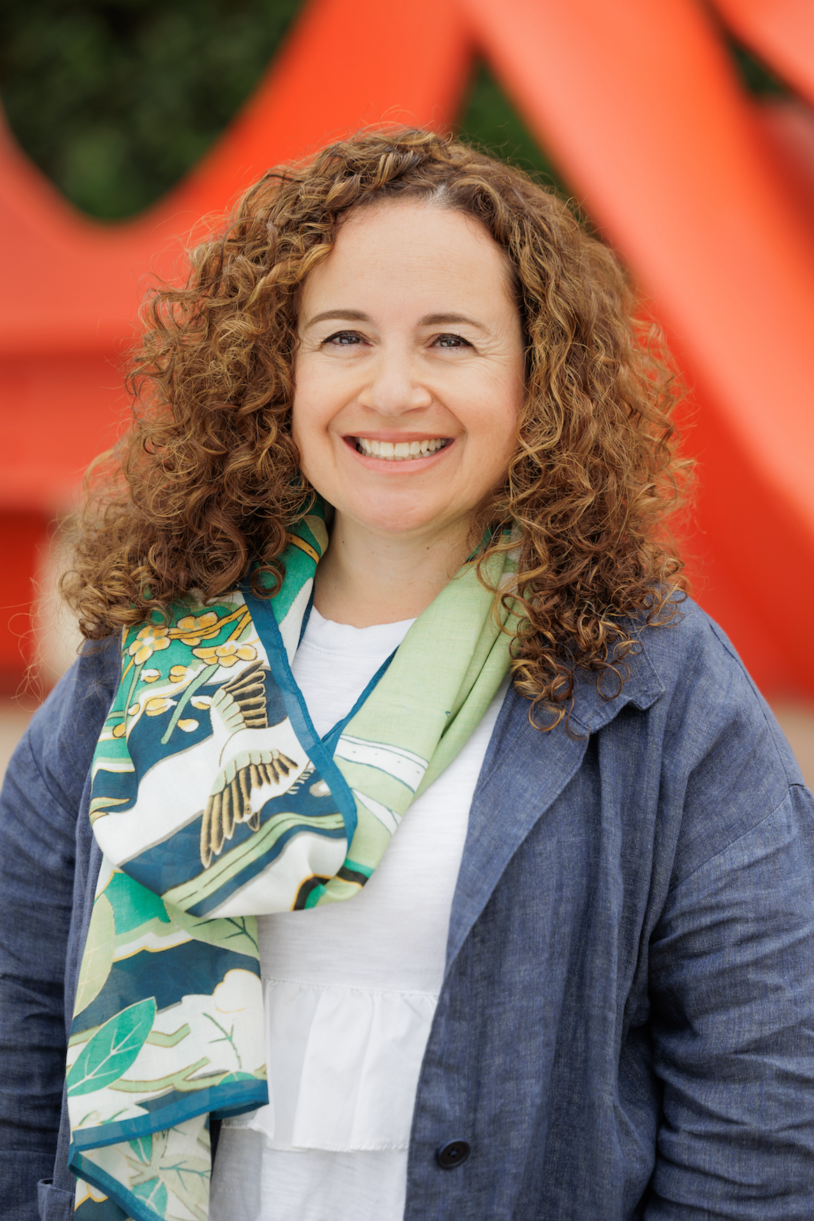 Image Description: A light-skinned woman with long curly hair stands outdoors, smiling broadly.