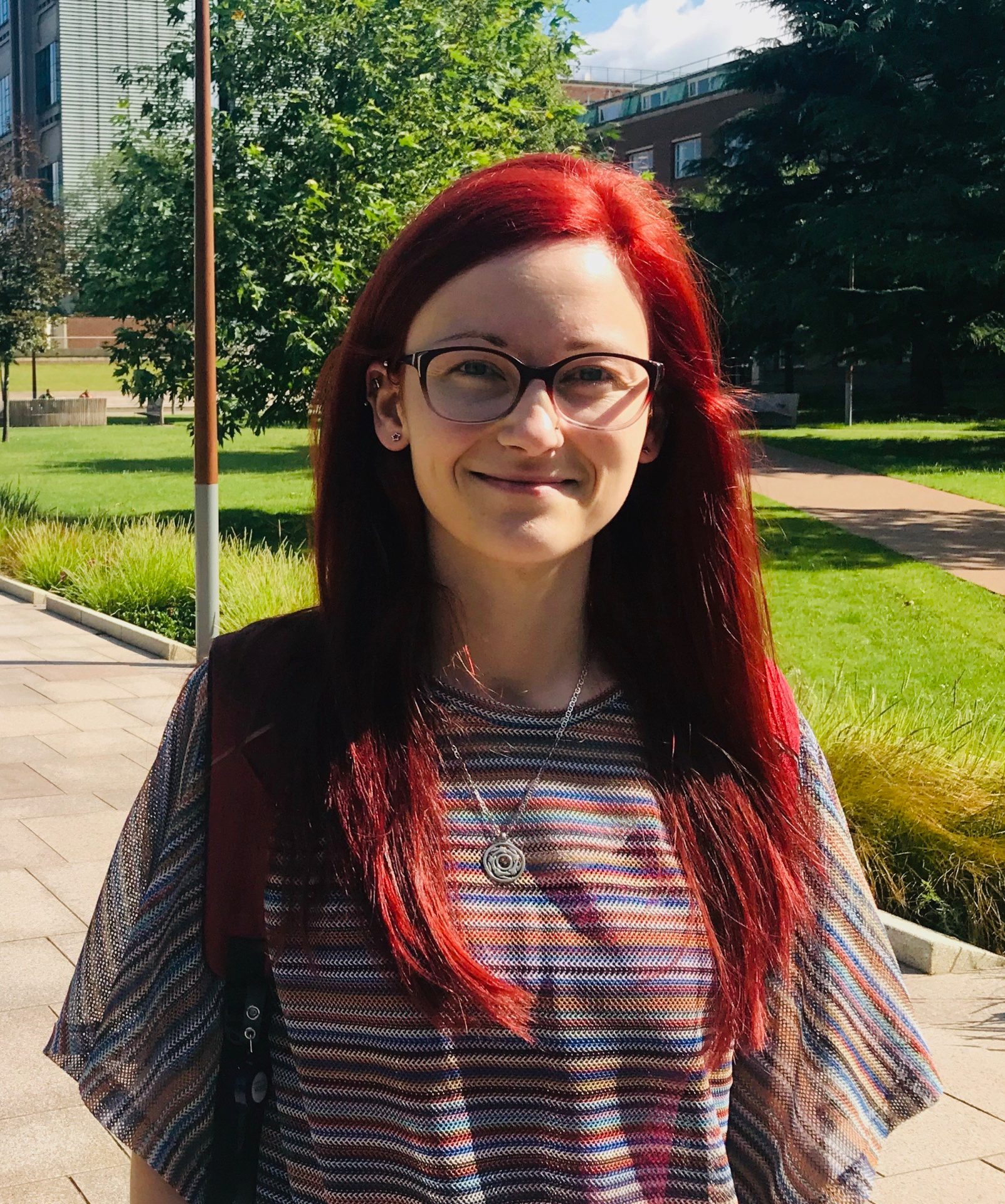 A light-skinned woman with long, straight, bright red hair stands outdoors, smiling at the camera. She wears glasses and a striped top.