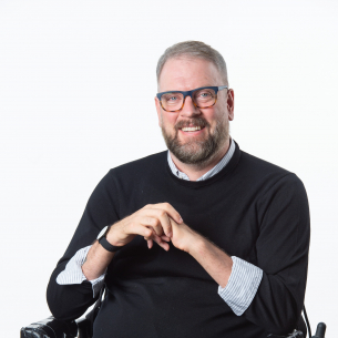 Image Description: A light-skinned man in a wheelchair wearing glasses and a black sweater over a pale blue button-down shirt.