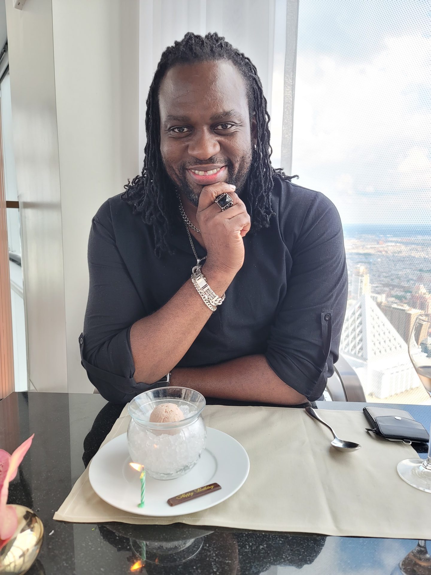 Image Description: A dark-skinned man with shoulder-length hair in small braids is seated at a table; he smiles at the camera. There is a dessert with a candle on the table in front of him, and through a window behind him, a city can be seen from high up.