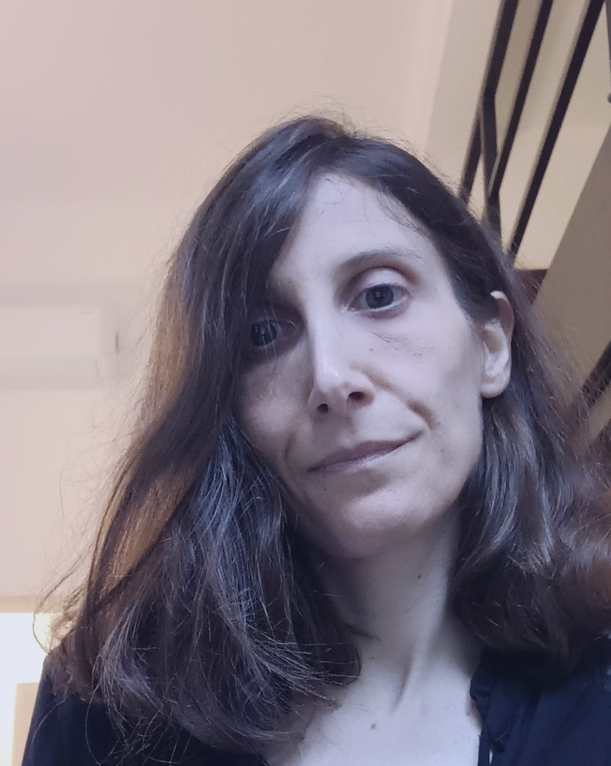 A pale woman with long brown hair stares pensively down and slightly away from the camera. She wears a black top.