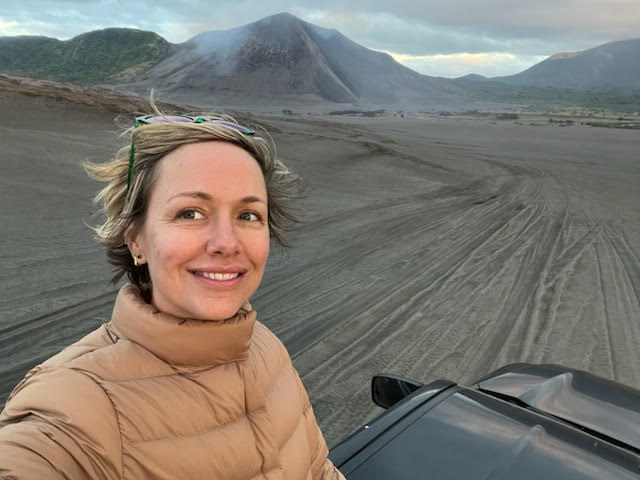 Image Description: A middle-age white woman with brown puffy coat smiling with smoldering volcano in the background.