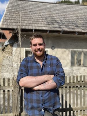 Image Description: portrait of a smiling fair-skinned person with short, sandy hair and beard, standing in front of a home with a fence in front. They wear a blue plaid button-down shirt.