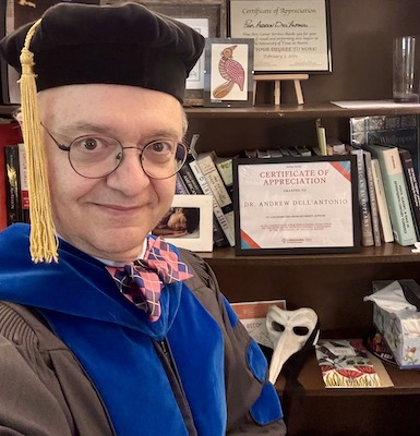 Image Description: Self portrait of a smiling light-skinned man wearing graduation cap and robes. Visible in the background are a number of awards and small art pieces on a bookshelf.