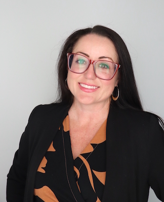 Image Description: portrait of a smiling light-skinned woman with long dark hair and glasses. She wears a black sweater over a printed top.