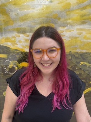 Image Description: portrait of a smiling, light-skinned person with long, straight, pink hair. They wear a black shirt and red-framed glasses.