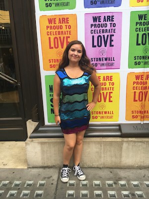 Image Description: A smiling, light-skinned femme person with long brown hair stands with one hand on their hip. They wear a short, sequined dress in shades of blue, with black sneakers. In the background is a vibrant graphic commemorating the 50th anniversary of Stonewall.
