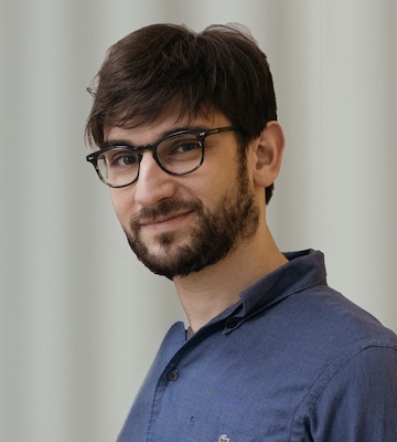 Image Description: portrait of a smiling, light-skinned man with medium-length brown hair and beard. He wears a blue button-down shirt and glasses.