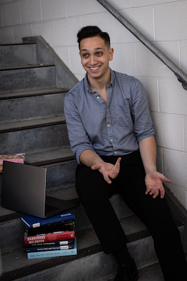 Image Description: a light brown skinned-man person short dark hair sits on a stairwell smiling broadly. Books are visible in the foreground, and one hand is open on their knee, as if for emphasis. They wear a blue button-down shirt and black slacks.