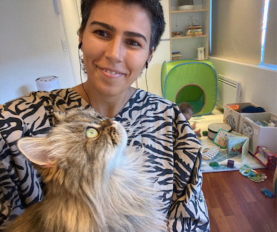 Image Description: a smiling, olive-skinned, short-haired woman sits holding a fluffy cat. She wears a printed shirt and headphones, and in the background a baby sits up and plays in a little area full of padding and enriching toys.