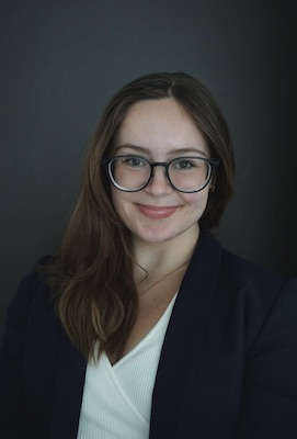 Image Description: portrait of a smiling fair-skinned woman with long brown hair. She wears glasses and a black blazer over a white top.
