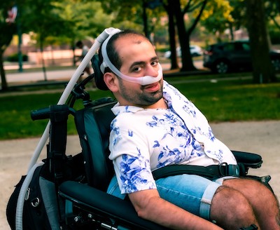 Image Description: a smiling, light-skinned man in a wheelchair. His dark hair is short, and he wears a printed button-down shirt and shorts.