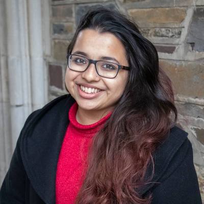 Image Description: Jiya, a femme-presenting South Asian person, wearing a black coat and red sweater, posed against a stone wall. They have long hair coming down their left shoulder, are wearing glasses and a nose-ring, and are smiling at the camera.