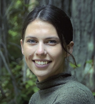 Image Description: portrait of a smiling, light-skinned feminine person with straight dark hair drawn back. In the background a tree and foliage can be seen.