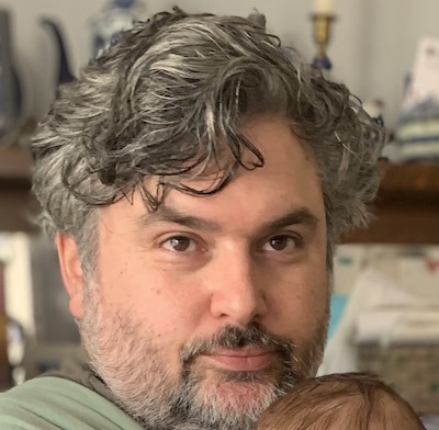 Image Description: portrait of a fair-skinned man with a thoughtful expression. His wavy, salt-and-pepper hair is swept back from his face, and his beard is cropped short.