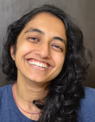 Image Description: portrait of a person with brown skin and long black hair smiling warmly, as if mid-laugh. They wear a blue top and a thin beaded necklace.