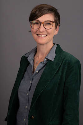 Image Description: Portrait of a smiling light-skinned woman with short, brown hair. She wears glasses, and a black blazer over a grey button-down shirt.