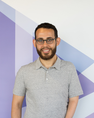 Image Description: portrait of a smiling olive-skinned man. His dark hair and beard are short; he wears glasses and a beige polo shirt.