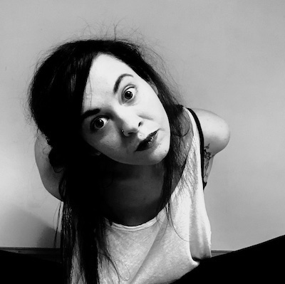 Image description: a black and white portrait of a light-skinned woman bent forward toward the camera, her head cocked to one side. Her expression is playful yet direct. Her long, dark, straight hair is pulled pack; she wears a white tank top.