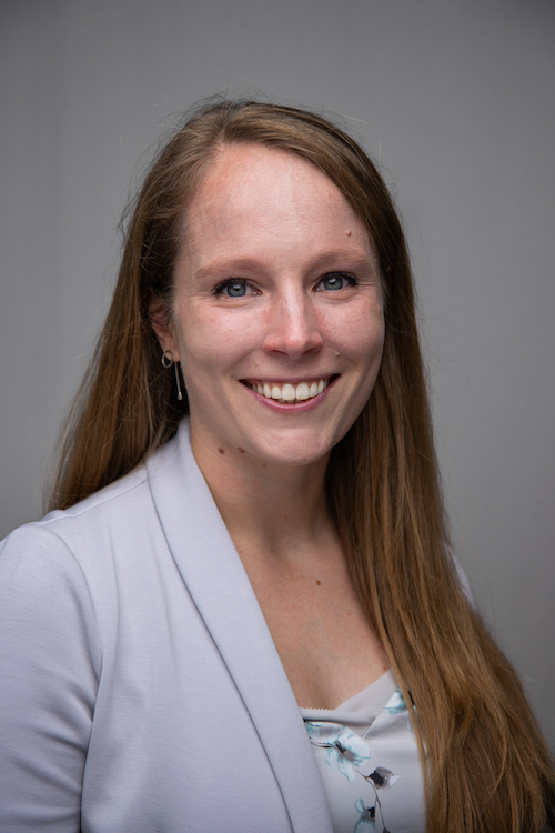 Image Description: portrait of a smiling fair-skinned woman with long red hair. She wears a white blazer over a printed top.