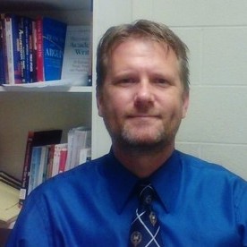 Image Description: portrait of a light-skinned man with short, sandy hair. He wears a blue button down shirt and tie; a bookshelf can be seen in the background.