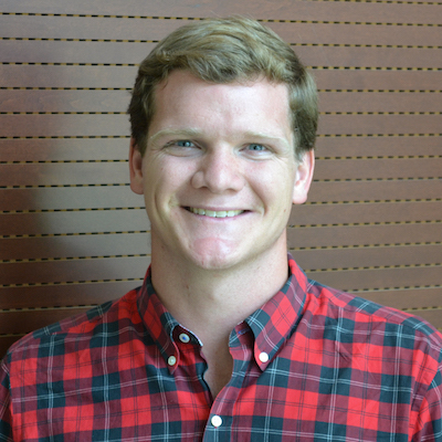 Image Description: portrait of a smiling, fair-skinned man with short blonde hair. He wears a button-down shirt with a red plaid pattern.