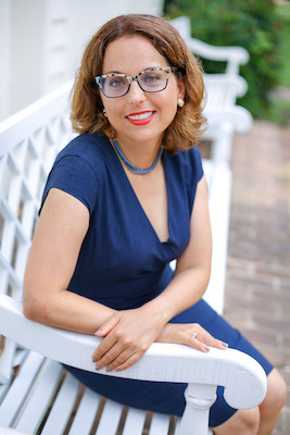 Image Description: A smiling light-skinned woman seated on a white bench. She wears a blue dress, necklace, and glasses; her brown hair is medium-length.