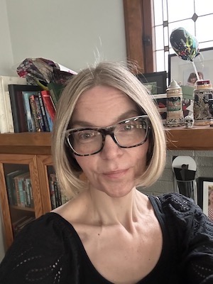 Image Description: A smiling, pale-skinned woman with chin-length, sandy hair. She wears glasses and a black top. In the background is visible a bookshelf and window.
