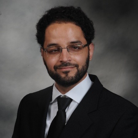 Image Description: portrait of a smiling, olive-skinned man with short black hair and beard. He is wearing a black suit and glasses.