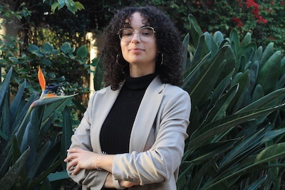 Image Description: portrait of a smiling light brown-skinned, femme presenting person standing outdoors with arms folded. They wear their dark curly hair to their shoulders, a nose piercing, glasses, and a beige blazer over a black shirt. A bird of paradise flower is visible to one side among the leafy background.