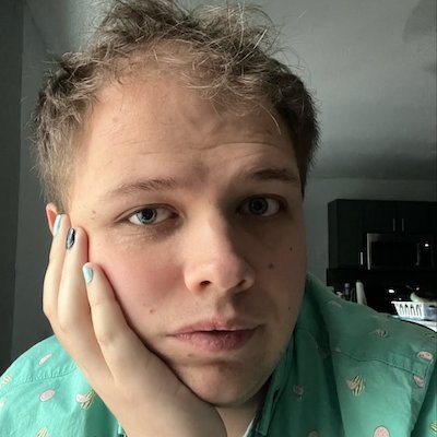Image Description: portrait of a fair-skinned person with a neutral expression and short, blonde hair. Their face rests in one hand, and their nails are painted multiple colors. They wear a printed green button-down shirt.