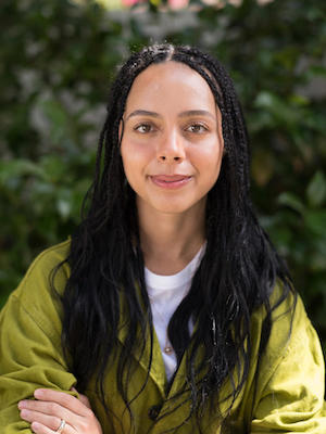 Image Description: portrait of a brown-skinned woman smiling; Her long black hair is worn down, and she wears a green top over a white shirt.